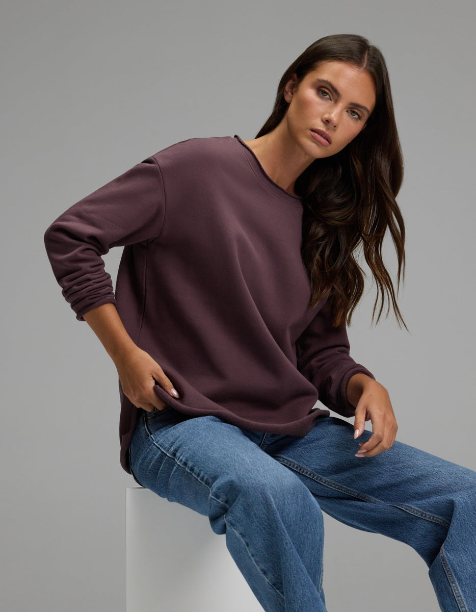 A woman with long brown hair sits on a white block against a plain gray background, looking at the camera with a relaxed expression while wearing the Cozy Earth Women’s Chelsea Pullover in maroon and blue jeans.