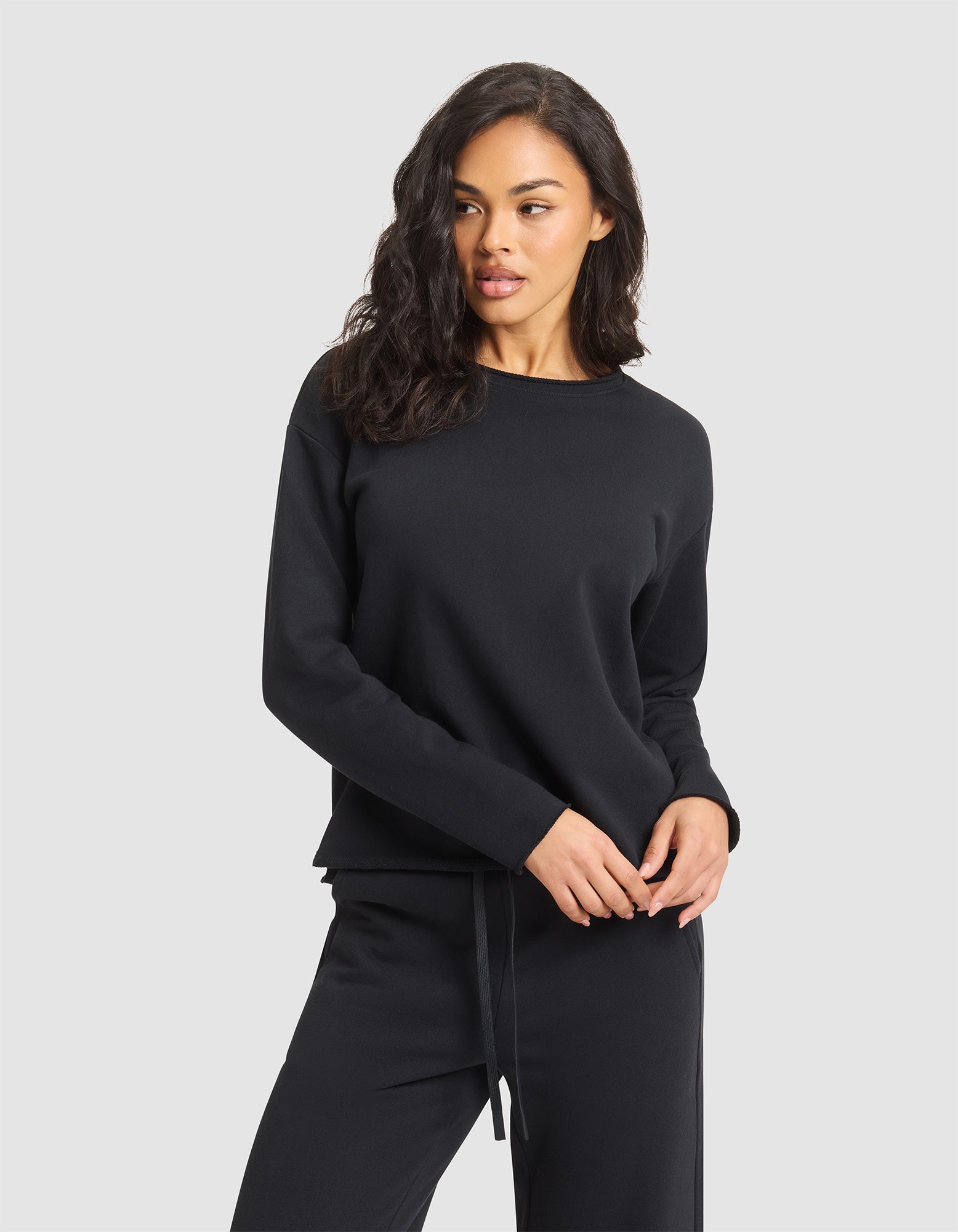 A woman with wavy dark hair models the Cozy Earth Women's Chelsea Pullover, a black long-sleeve sweatshirt, standing against a plain light gray background and looking slightly to her left. 