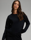 A woman with long brown hair is wearing the Cozy Earth Women’s Chelsea Pullover in Jet Black, paired with black pants. She stands with one hand on her hip, looking to the side against a plain gray background.|Color:Jet Black