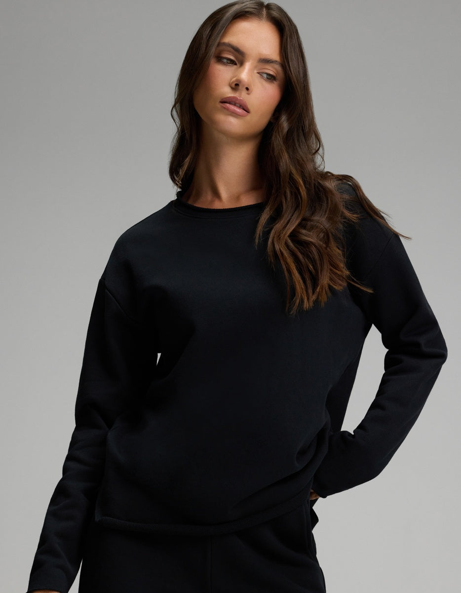 A woman with long brown hair is wearing the Cozy Earth Women’s Chelsea Pullover in Jet Black, paired with black pants. She stands with one hand on her hip, looking to the side against a plain gray background.|Color:Jet Black
