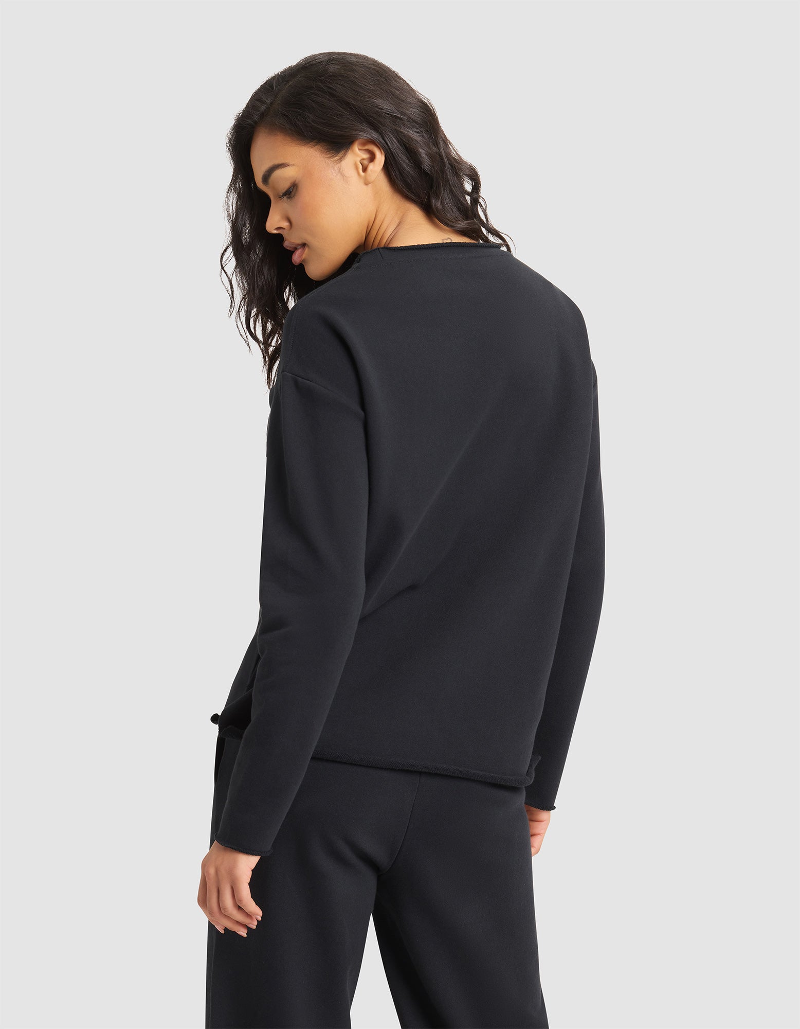 A woman with long, wavy hair faces away, looking over her shoulder. She wears the black Women's Chelsea Pullover by Cozy Earth with matching pants against a plain light gray background. 