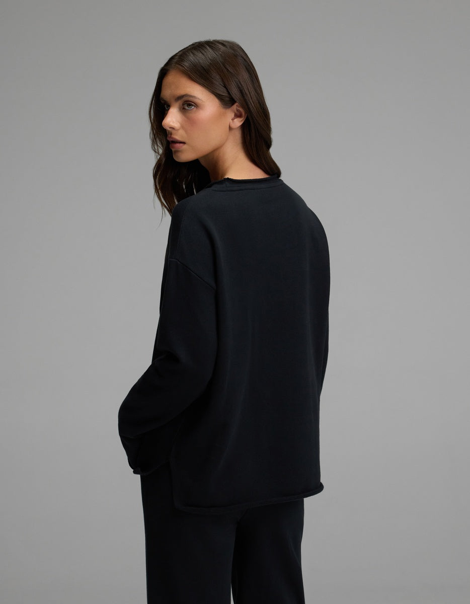 A woman with long brown hair stands sideways against a plain gray background, wearing the Cozy Earth Women’s Chelsea Pullover in black with her hands in her pockets.