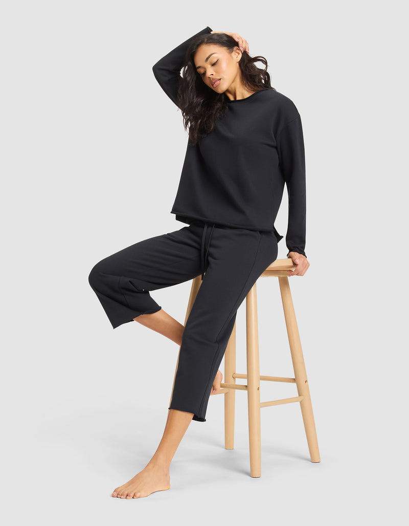A woman models the Cozy Earth Women's Chelsea Pullover, a loose black long-sleeve top, paired with cropped pants. She sits barefoot on a wooden stool, one hand resting on her head, against a plain light gray background.