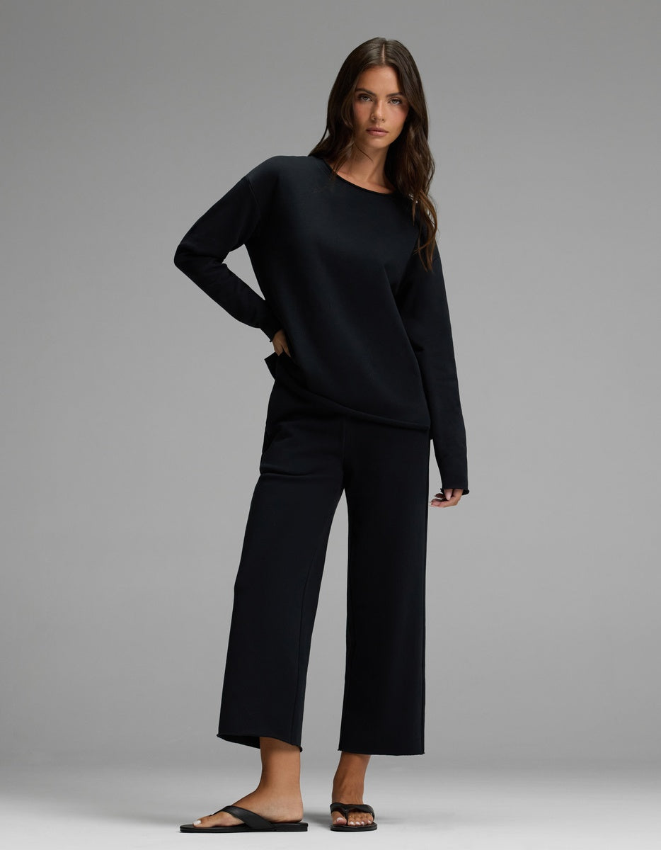 A woman with long brown hair and a relaxed expression poses against a gray backdrop, wearing the Cozy Earth Women’s Chelsea Pullover in black, paired with wide-leg cropped pants and black sandals. She has one hand in her pocket.