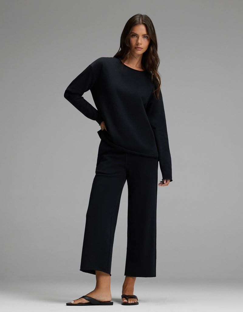 A woman with long brown hair and a relaxed expression poses against a gray backdrop, wearing the Cozy Earth Women’s Chelsea Pullover in black, paired with wide-leg cropped pants and black sandals. She has one hand in her pocket.