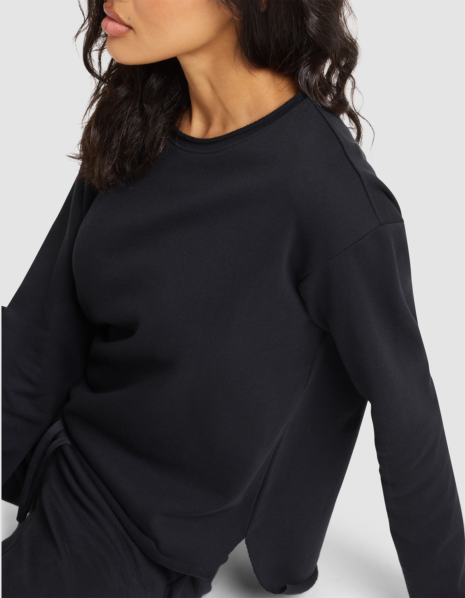 A woman with wavy dark hair, wearing the Cozy Earth Women's Chelsea Pullover in black with matching pants, sits on a light surface and faces slightly away from the camera. 