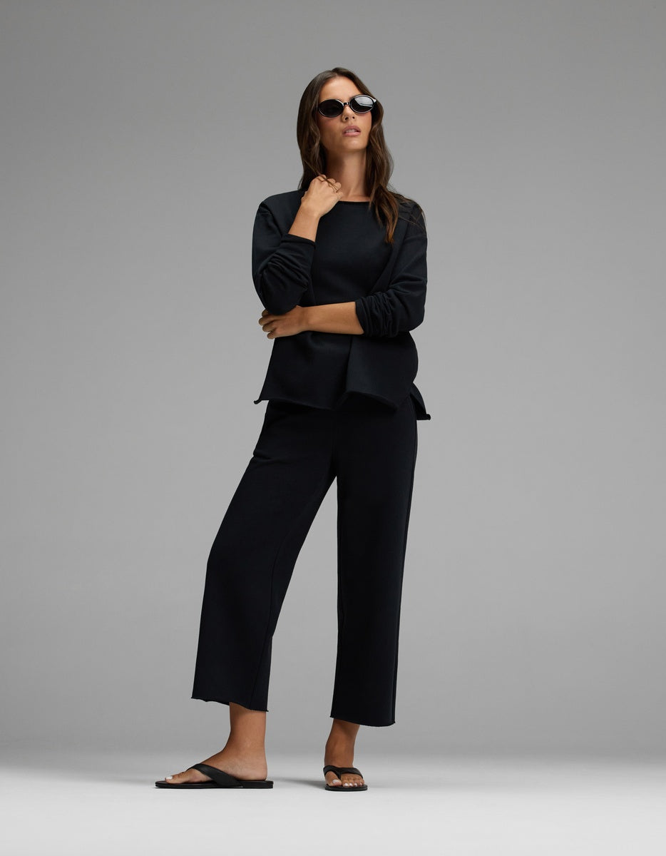 A woman stands confidently against a plain gray background, wearing the Cozy Earth Women’s Chelsea Pullover in black, paired with black wide-leg pants, black sunglasses, and black sandals.