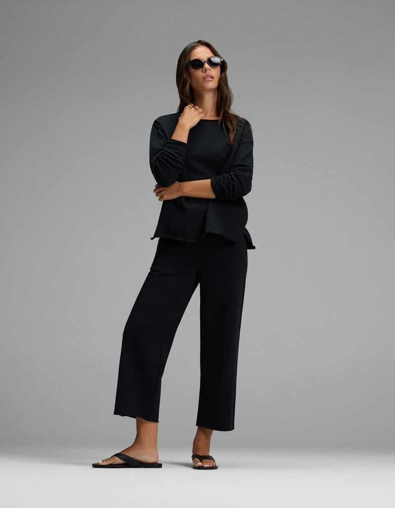 A woman stands confidently against a plain gray background, wearing the Cozy Earth Women’s Chelsea Pullover in black, paired with black wide-leg pants, black sunglasses, and black sandals.