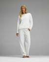 A woman with long blonde hair stands barefoot against a plain gray background, wearing the Women’s Chelsea Set— a loose-fitting white long-sleeve top and matching white pants from Cozy Earth.|Color:Bone