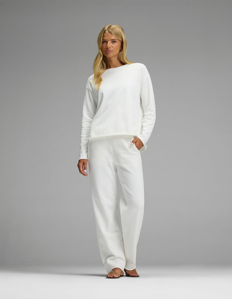 A woman with long blonde hair stands barefoot against a plain gray background, wearing the Women’s Chelsea Set— a loose-fitting white long-sleeve top and matching white pants from Cozy Earth.