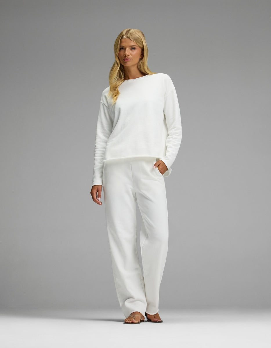 A woman with long blonde hair stands barefoot against a plain gray background, wearing the Women’s Chelsea Set— a loose-fitting white long-sleeve top and matching white pants from Cozy Earth.|Color:Bone