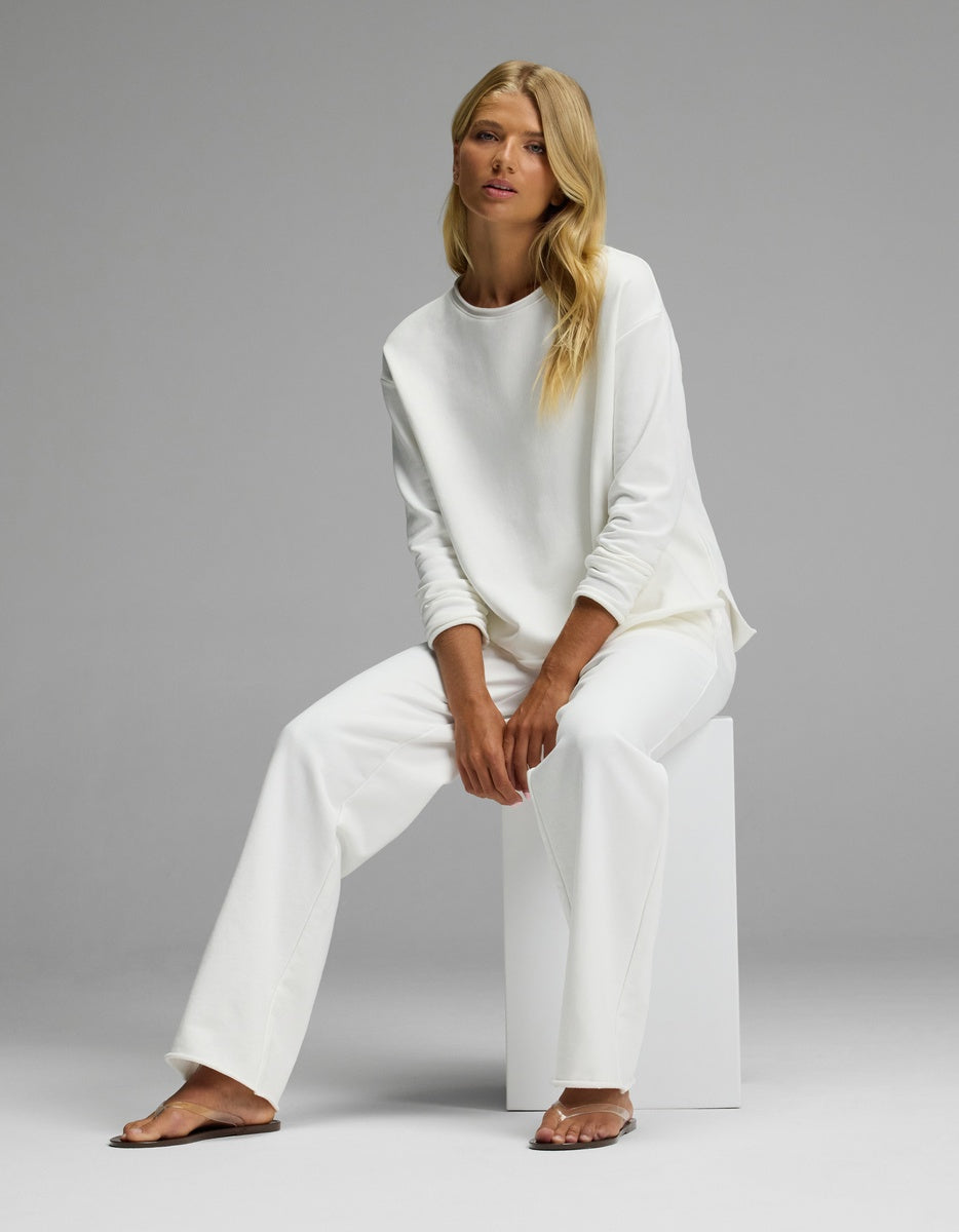 A woman with long blonde hair sits on a white block, wearing the Women’s Chelsea Set by Cozy Earth—a loose-fitting white long-sleeve top and matching white pants—against a plain gray background.
