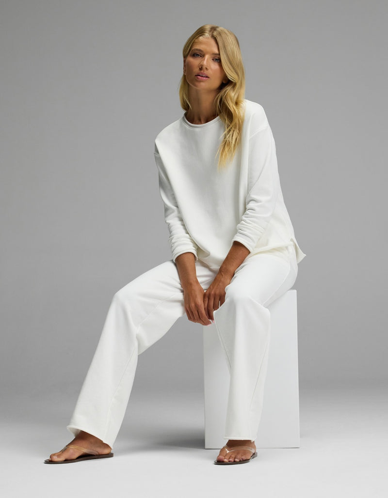 A woman with long blonde hair sits on a white block, wearing the Women’s Chelsea Set by Cozy Earth—a loose-fitting white long-sleeve top and matching white pants—against a plain gray background.