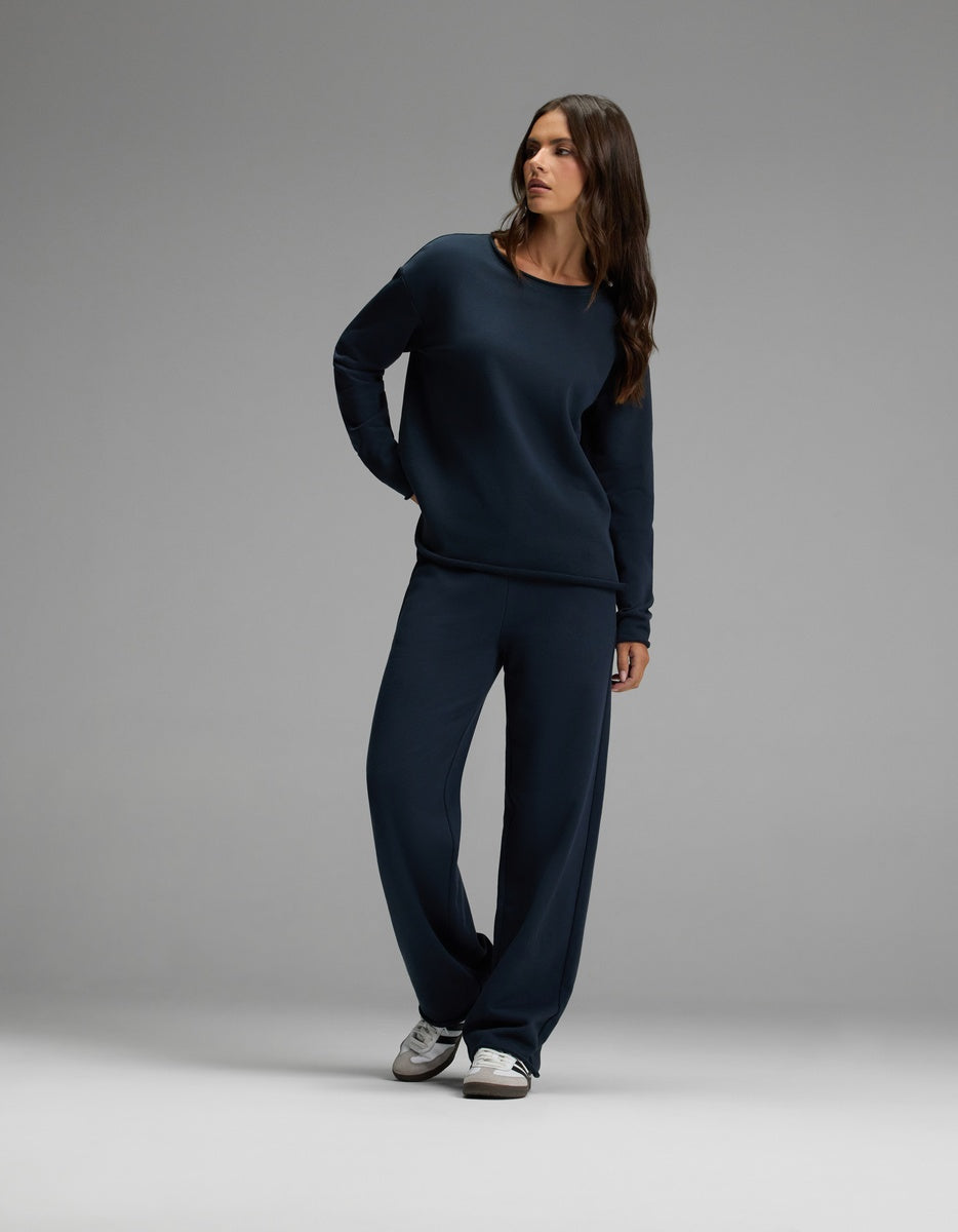 A woman wearing the Cozy Earth Women’s Chelsea Set in Eclipse—featuring a dark blue long-sleeve top and matching wide-leg pants—stands against a plain gray background, looking to the side. She has long brown hair and is wearing white sneakers.