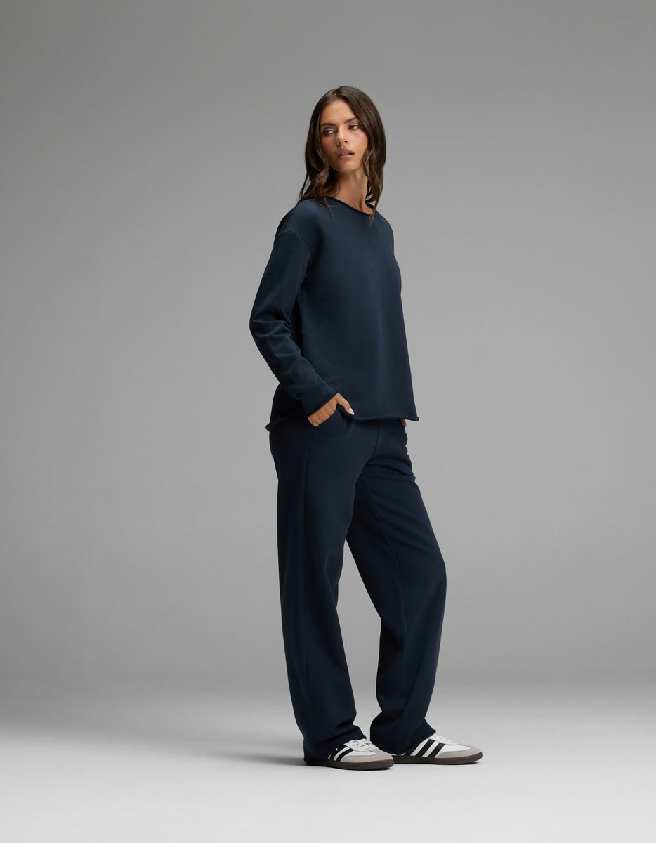 A person wearing the Women’s Chelsea Set by Cozy Earth—a loose-fitting navy sweatshirt and matching pants—paired with white Adidas sneakers stands against a plain gray backdrop, looking slightly to the side.