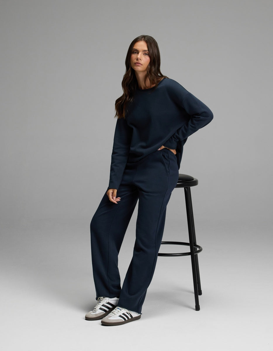 A woman with long brown hair wears the Cozy Earth Women’s Chelsea Set in navy blue, standing with one hand in her pocket and the other on a black stool. She has a neutral expression and is wearing white Adidas sneakers.