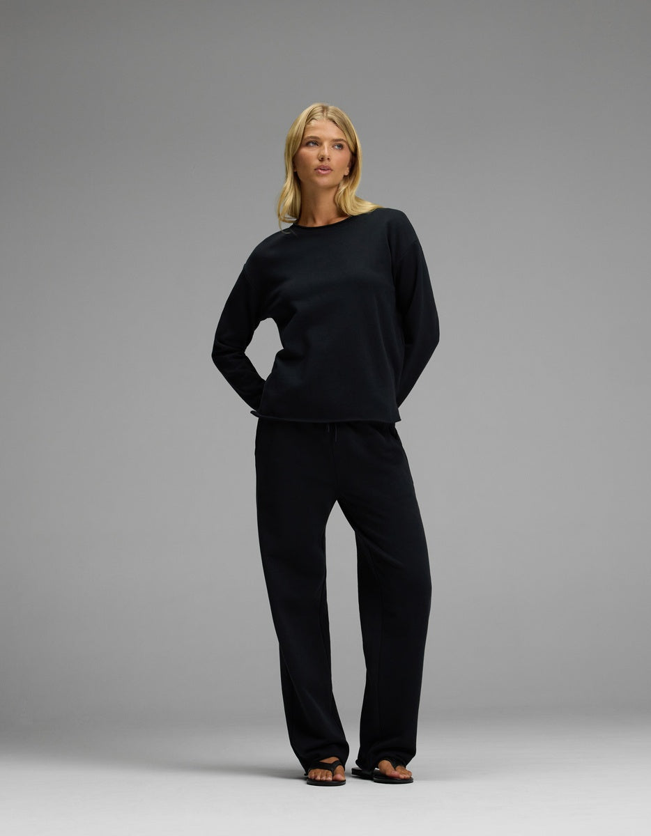 A woman stands against a gray background, wearing the Cozy Earth Women’s Chelsea Set—a loose-fitting black long-sleeve top paired with matching black pants—and black sandals. Her shoulder-length blonde hair frames her face as she rests her hands behind her back and gazes to the side.|Color:Jet Black