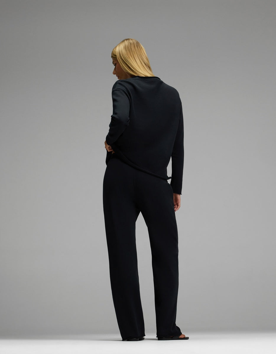 A person with long blonde hair stands barefoot facing away against a plain gray background, wearing the Cozy Earth Women’s Chelsea Set—a loose black long-sleeve top and matching black pants.