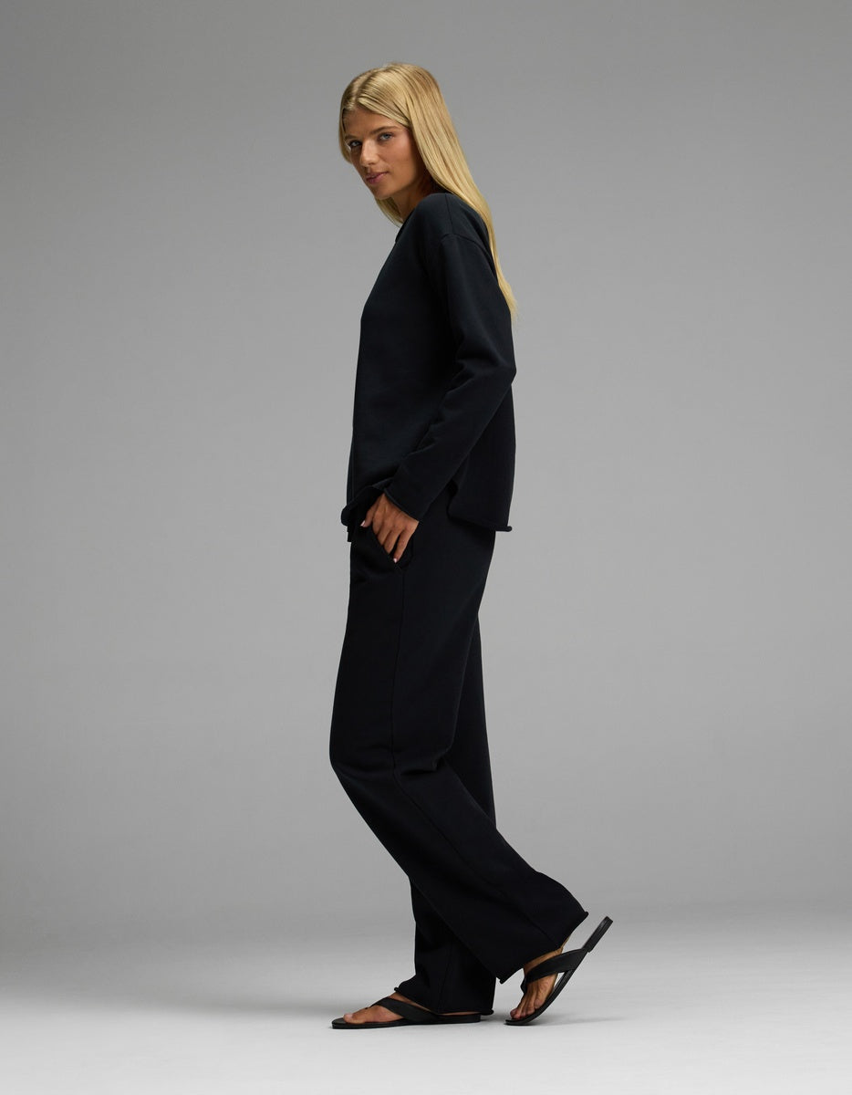 A person with long blonde hair, wearing the Cozy Earth Women’s Chelsea Set—featuring a loose black long-sleeve top and matching black pants—and black sandals, stands sideways against a plain gray background.