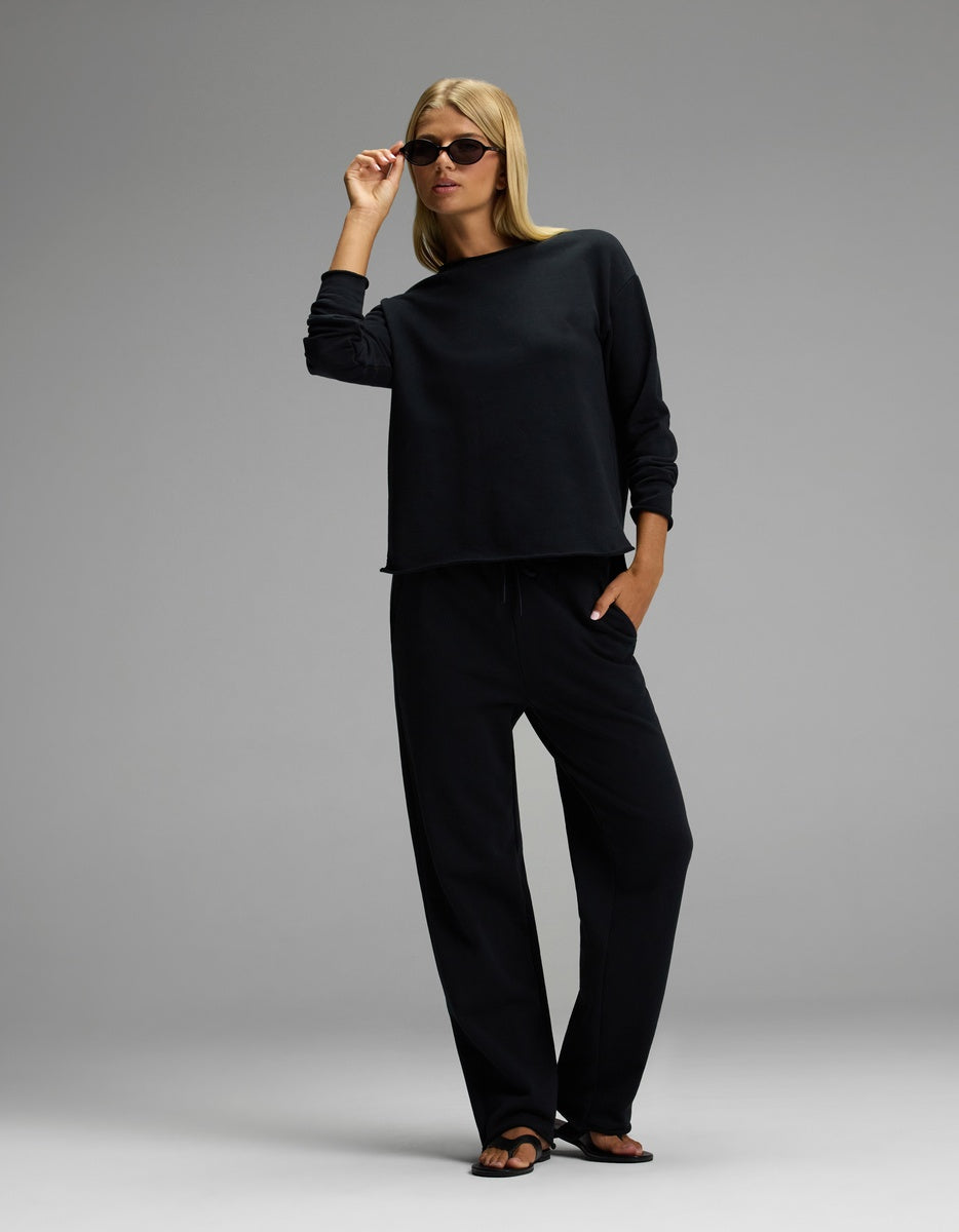 A woman wearing the Cozy Earth Women’s Chelsea Set—a loose black long-sleeve top and matching black pants—stands against a plain gray background with one hand in her pocket and the other adjusting her sunglasses.
