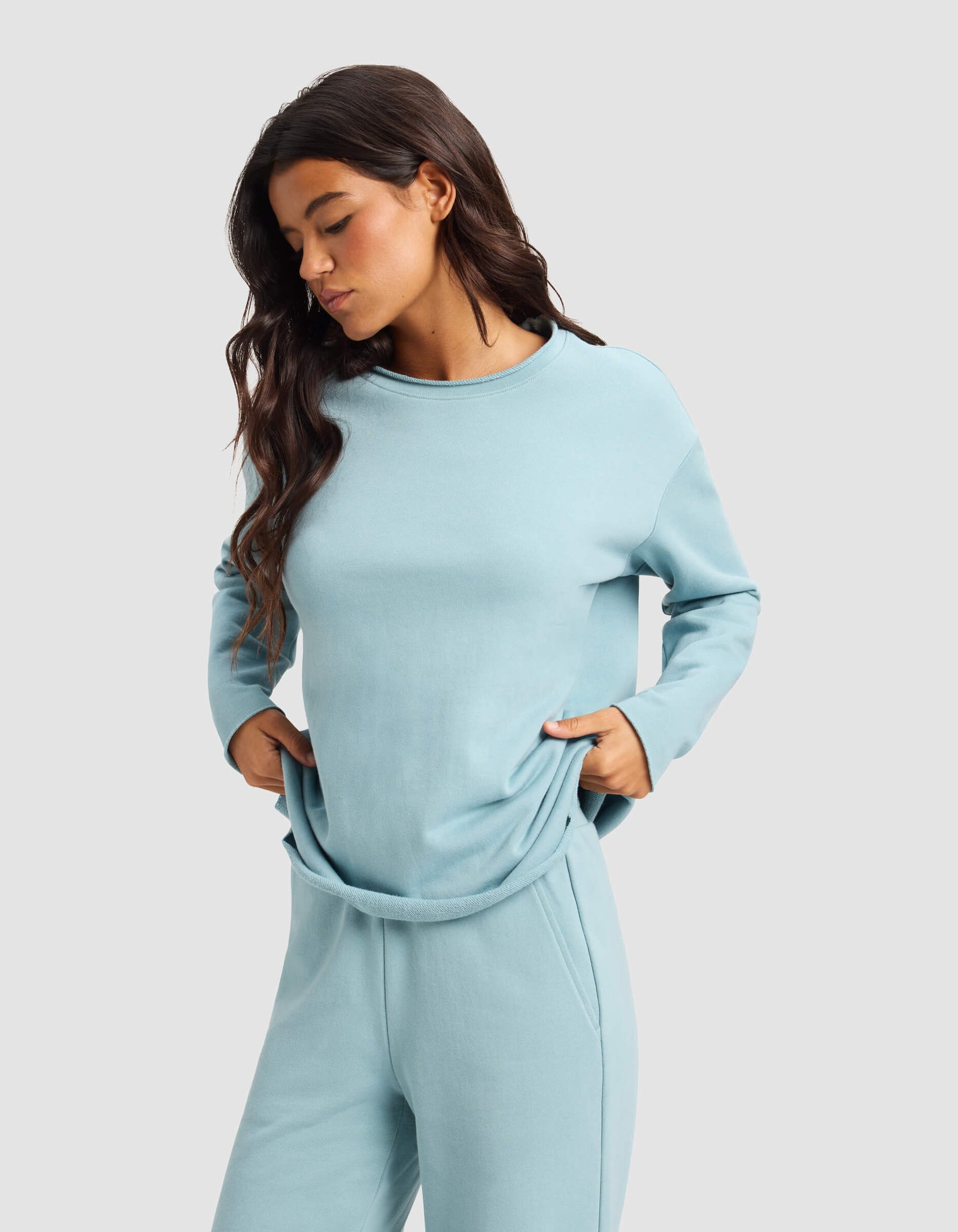 A woman with long brown hair wears the Cozy Earth Women's Chelsea Pullover in light blue and matching sweatpants, standing against a plain light gray background with her hands in her pockets, looking down.