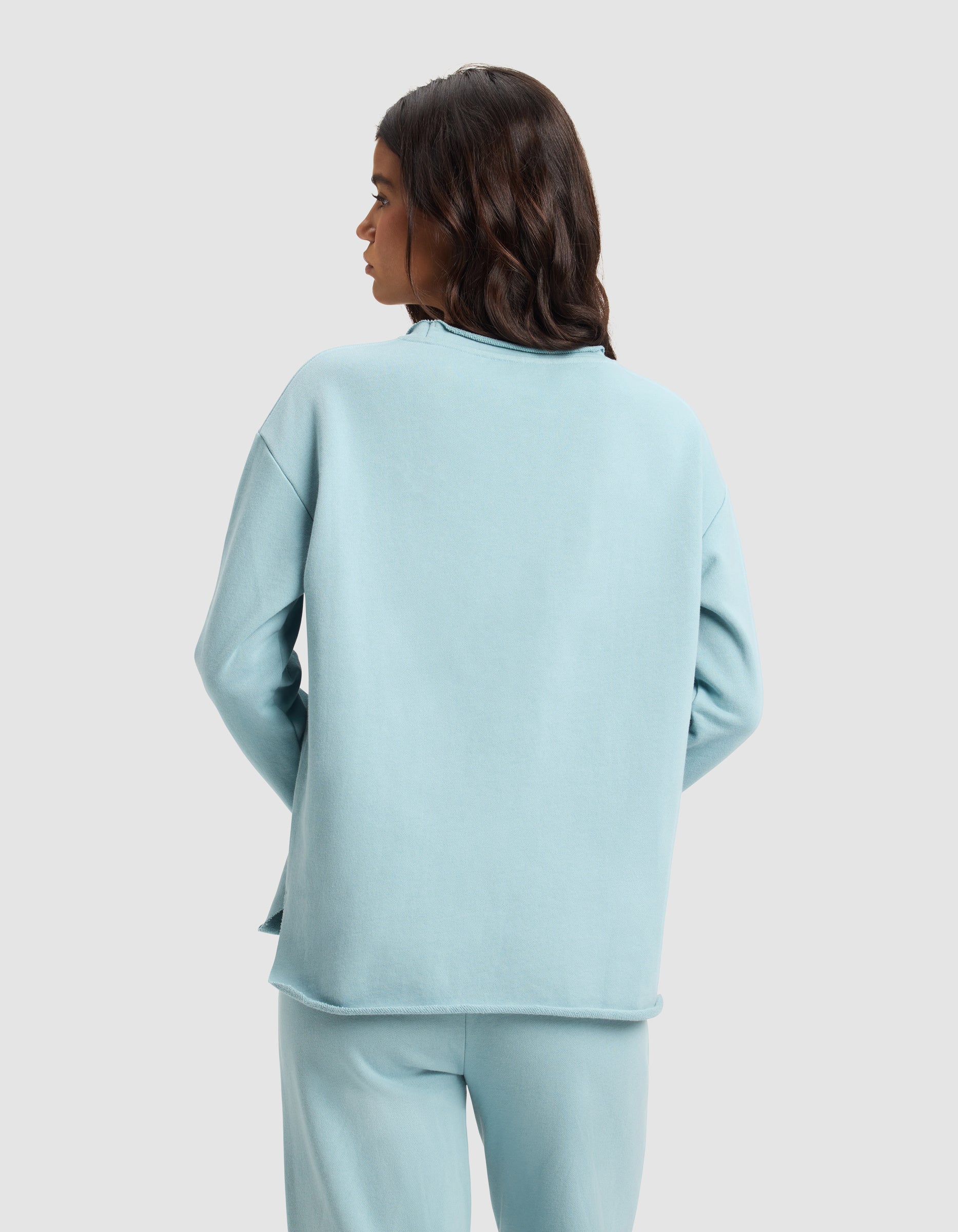 A woman with long, wavy dark hair is seen from behind wearing the Cozy Earth Women's Chelsea Pullover in light blue against a plain backdrop.|Color:River