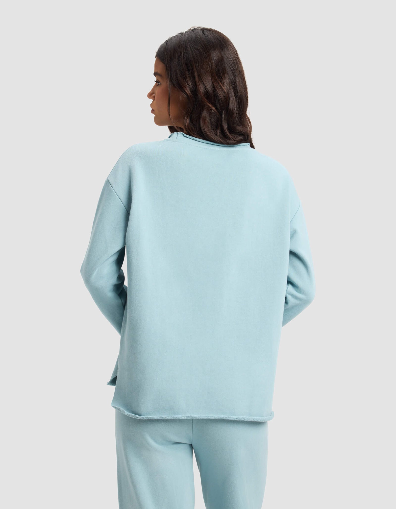 A woman with long, wavy dark hair is seen from behind wearing the Cozy Earth Women's Chelsea Pullover in light blue against a plain backdrop.