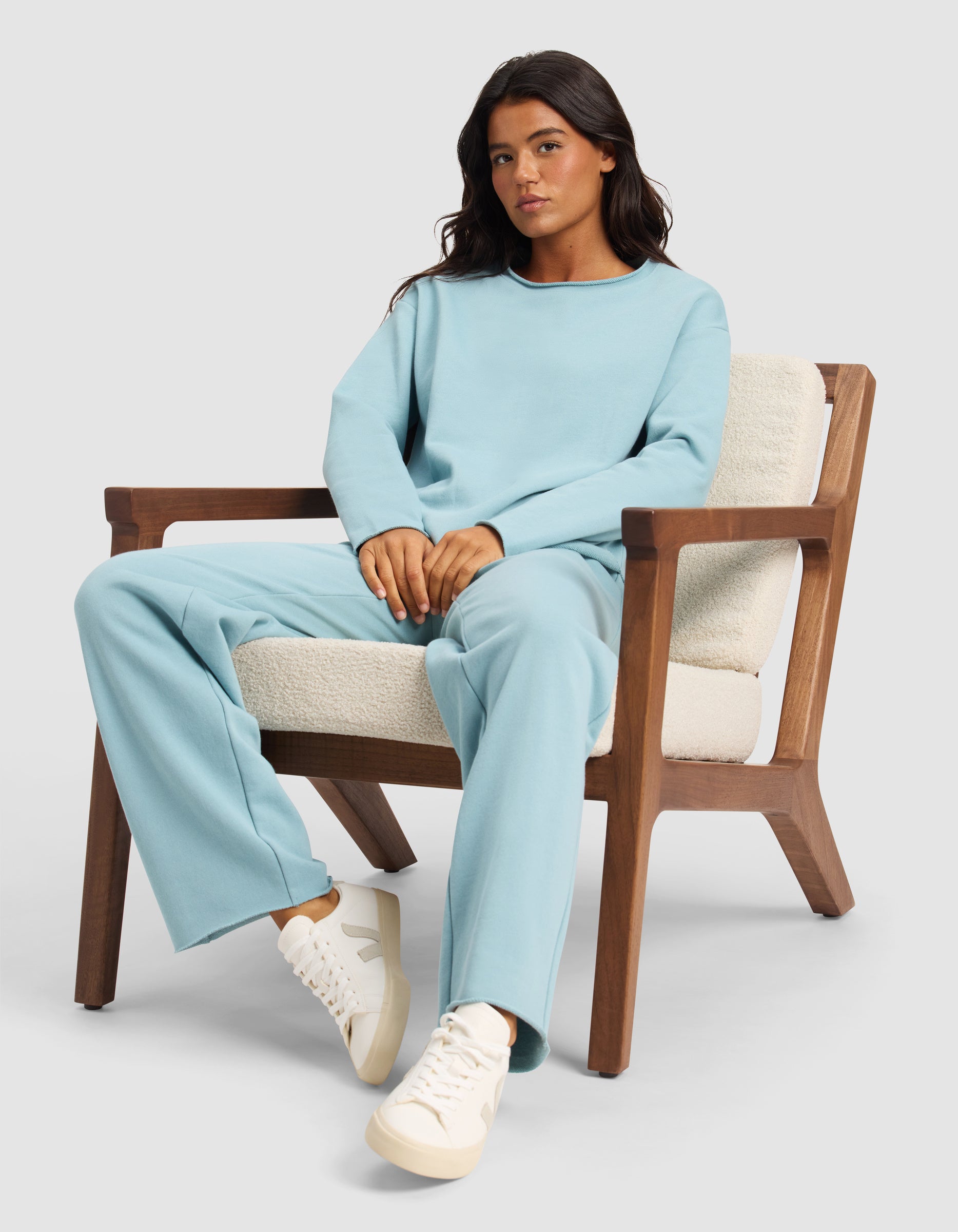 A woman sits calmly in a wooden armchair with cream cushions, wearing the Cozy Earth Women's Chelsea Pullover in light blue and white sneakers, looking at the camera against a plain light background.|Color:River