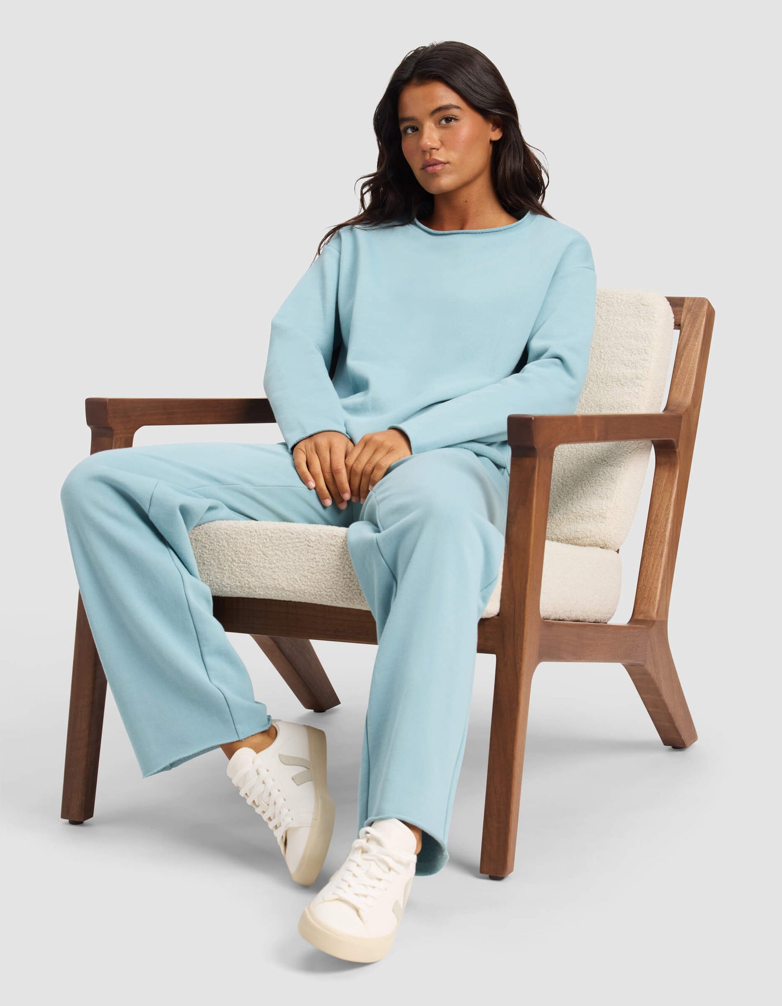 A woman sits calmly in a wooden armchair with cream cushions, wearing the Cozy Earth Women's Chelsea Pullover in light blue and white sneakers, looking at the camera against a plain light background.