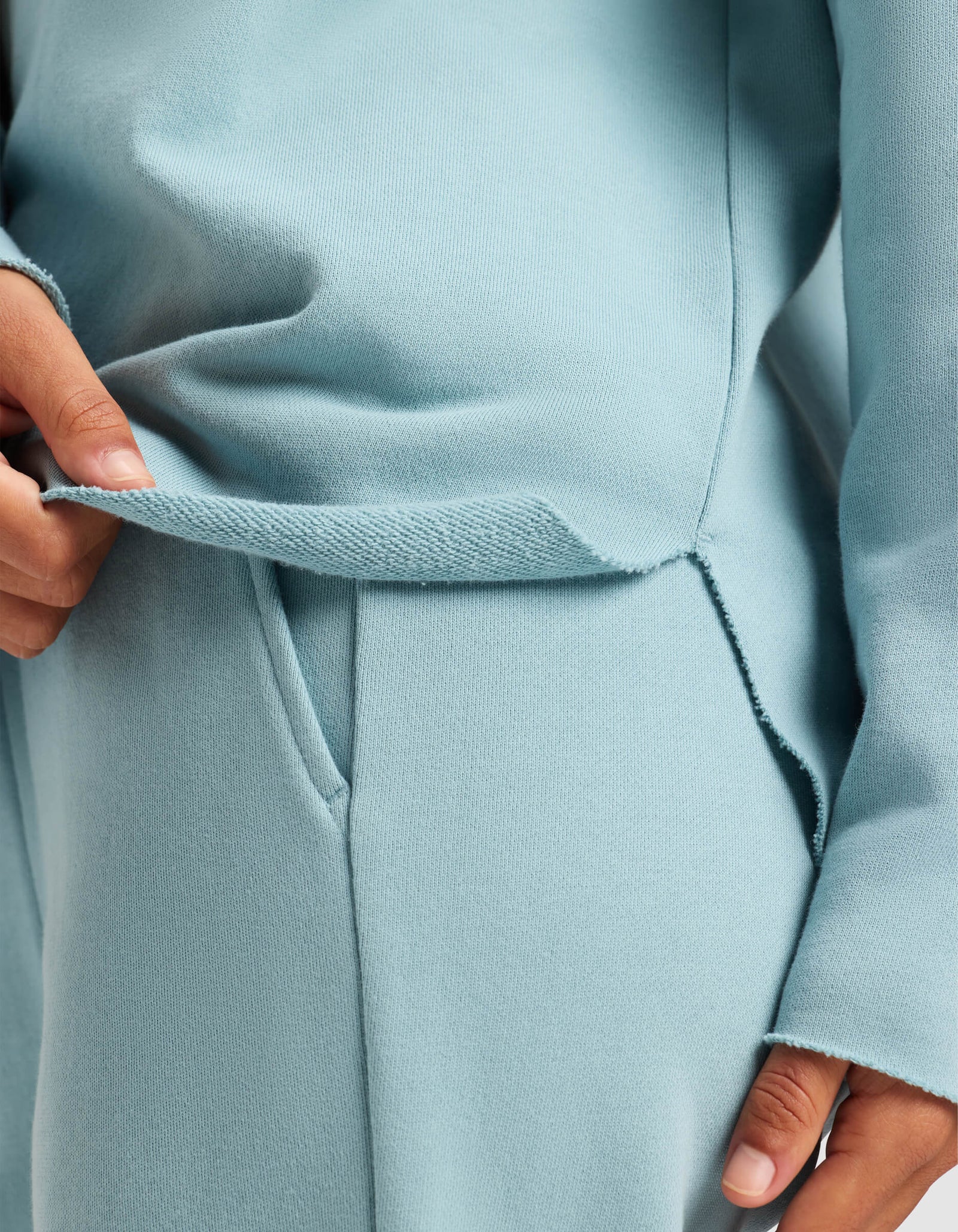A woman wears Cozy Earth’s Women’s Chelsea Pullover in light blue, lightly lifting the hem to reveal the side slit and soft fabric. The pullover’s relaxed fit highlights comfort and style.