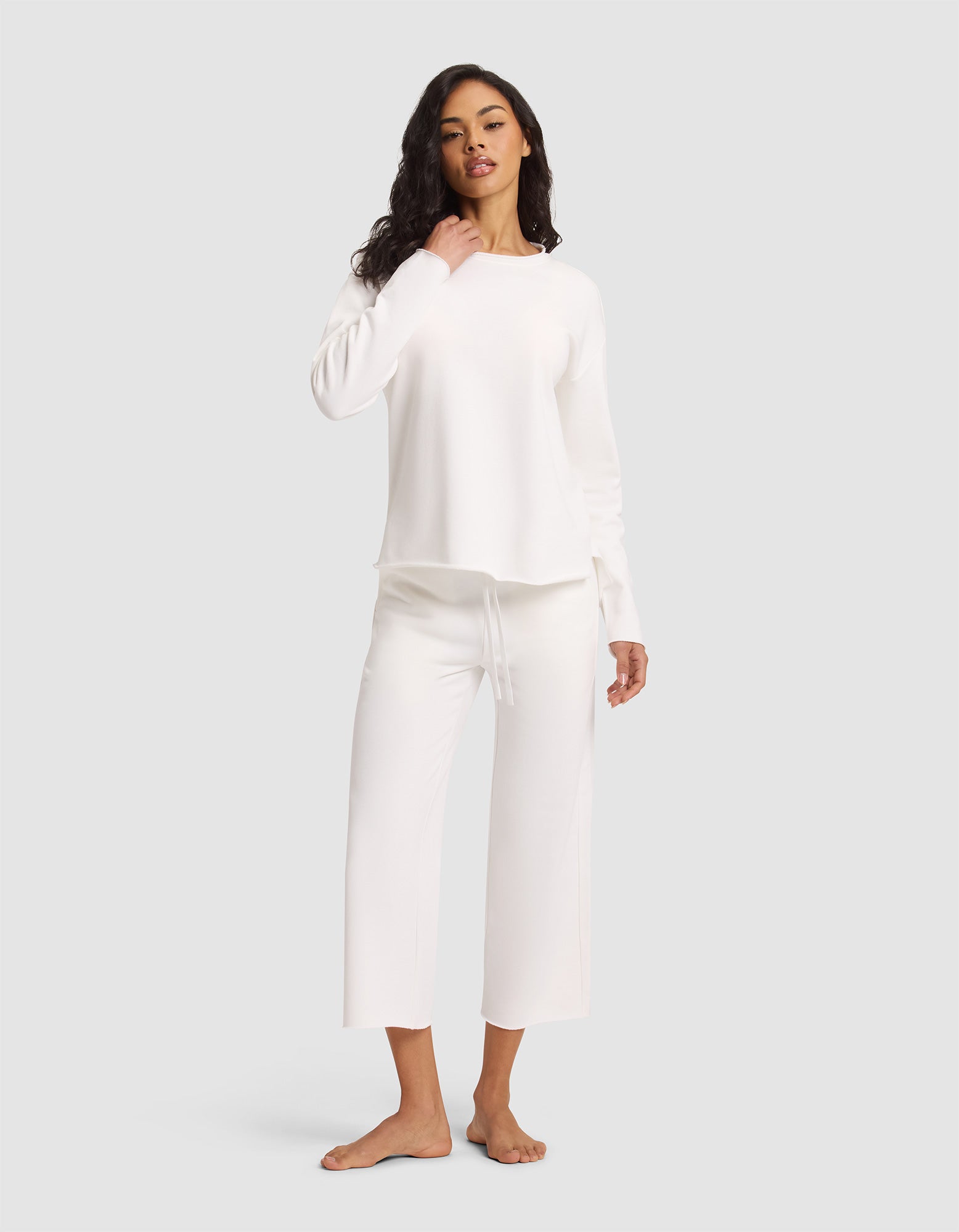 A woman poses barefoot against a plain backdrop, wearing Cozy Earth's Women's Chelsea Set—a long-sleeved white top and matching wide-leg drawstring pants—with one hand near her neck. 