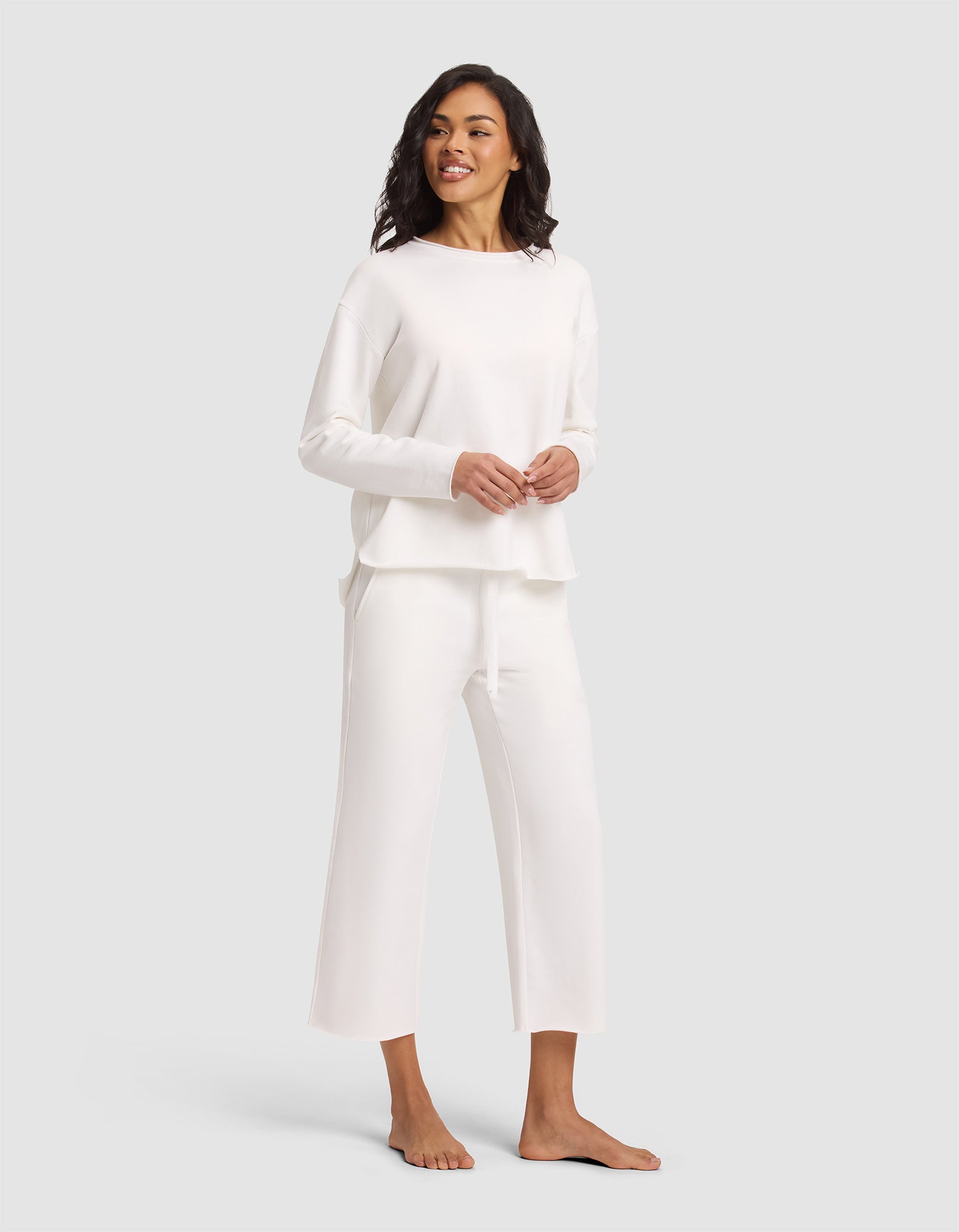 A smiling woman stands barefoot against a light gray background, wearing the Cozy Earth Women's Chelsea Set—loose-fitting, long-sleeved white top and matching wide-leg pants. 
