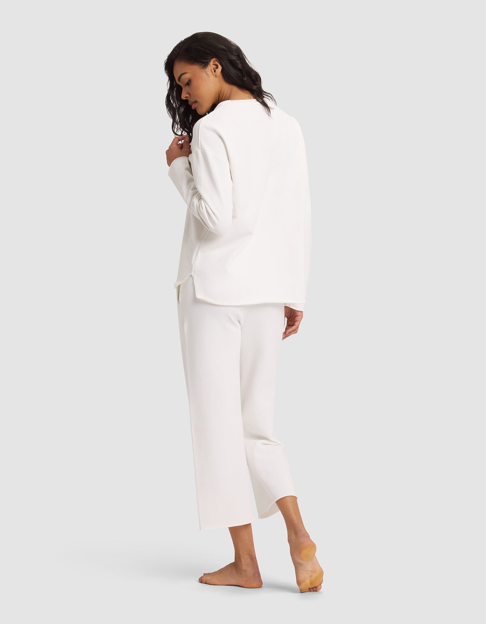 A woman with wavy dark hair, barefoot and looking over her shoulder, models the Women's Chelsea Set by Cozy Earth—featuring a loose-fitting long-sleeve white top and matching cropped pants—against a plain light gray background. 