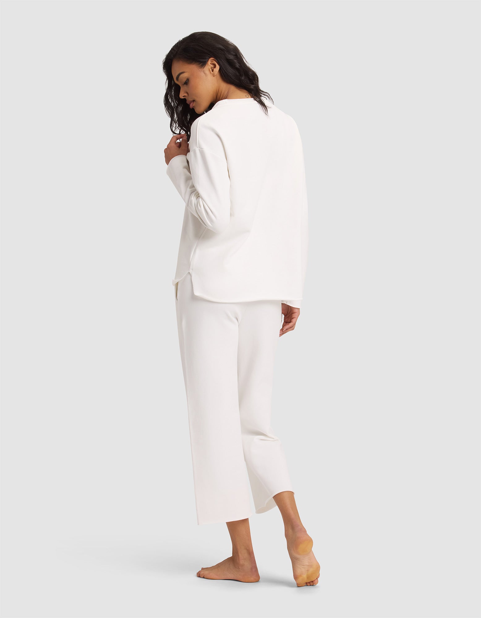 A woman with wavy dark hair, barefoot and looking over her shoulder, models the Women's Chelsea Set by Cozy Earth—featuring a loose-fitting long-sleeve white top and matching cropped pants—against a plain light gray background. 