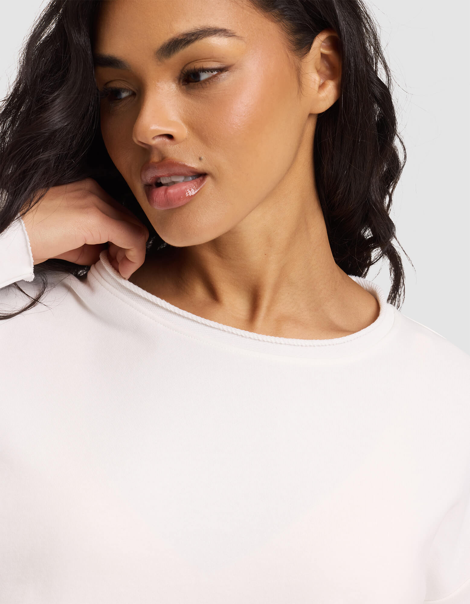 A woman with wavy dark hair and glowing skin, wearing the Cozy Earth Women's Chelsea Set, looks to the side while gently touching her neck. She is dressed in white against a neutral background. 