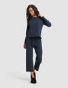 A woman with curly hair smiles and looks down while wearing Cozy Earth's Women's Chelsea Set—a navy blue long-sleeve top with matching wide-leg pants. She stands against a light gray background and wears black slippers.  |Color:Eclipse