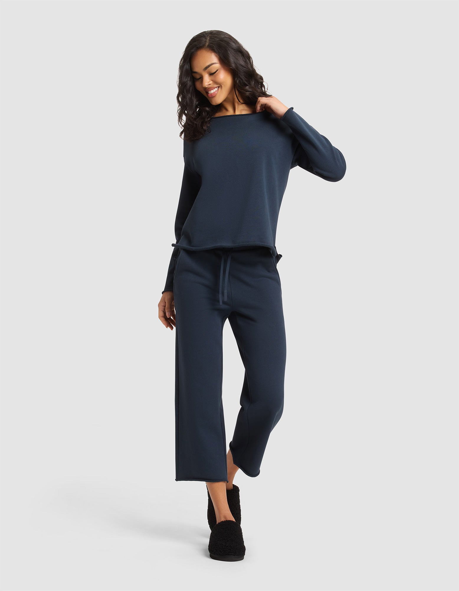 A woman with curly hair smiles and looks down while wearing Cozy Earth's Women's Chelsea Set—a navy blue long-sleeve top with matching wide-leg pants. She stands against a light gray background and wears black slippers.  |Color:Eclipse