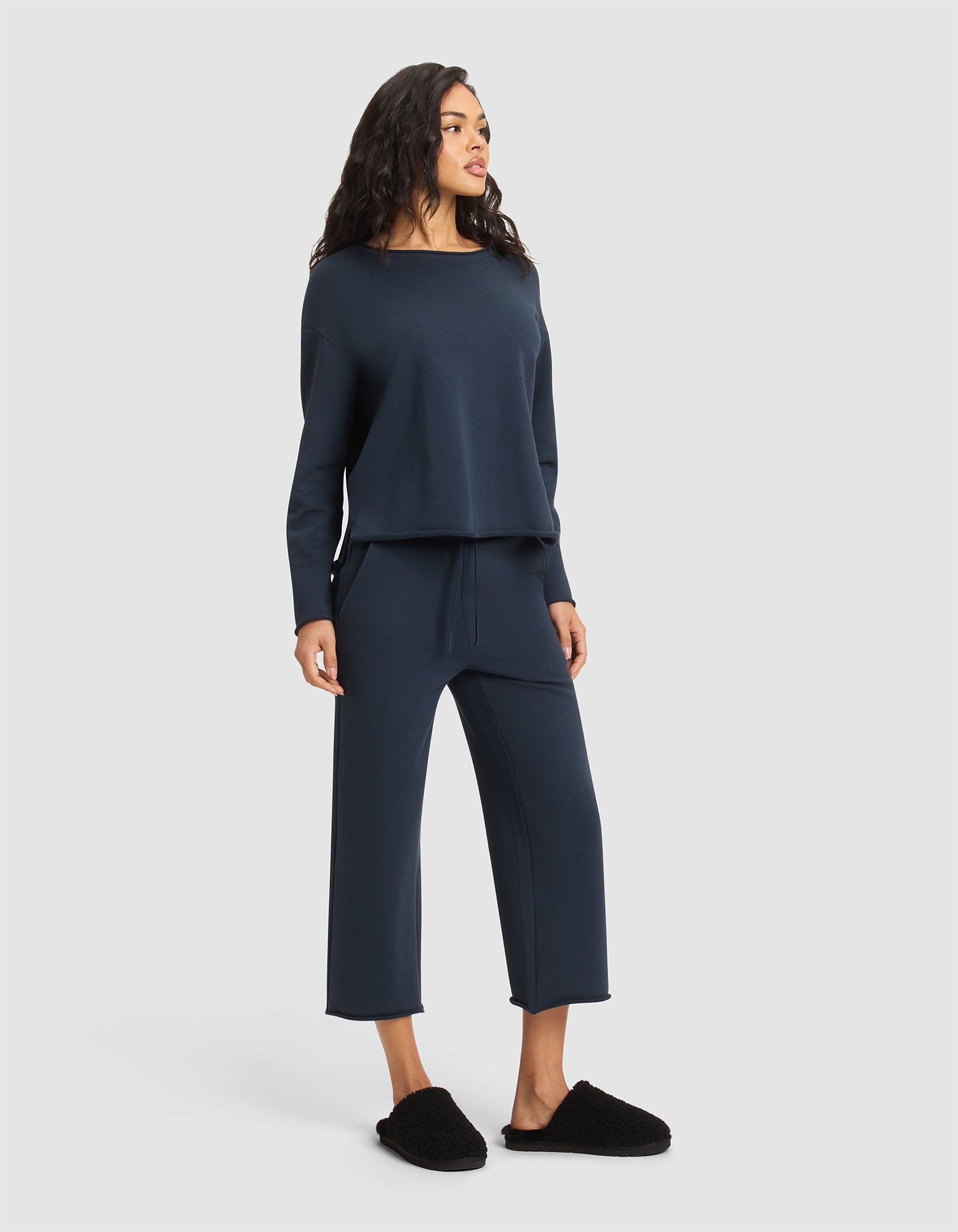 A woman stands against a plain background wearing the Cozy Earth Women's Chelsea Set in navy blue and black slippers, looking slightly to the side with a relaxed posture. 