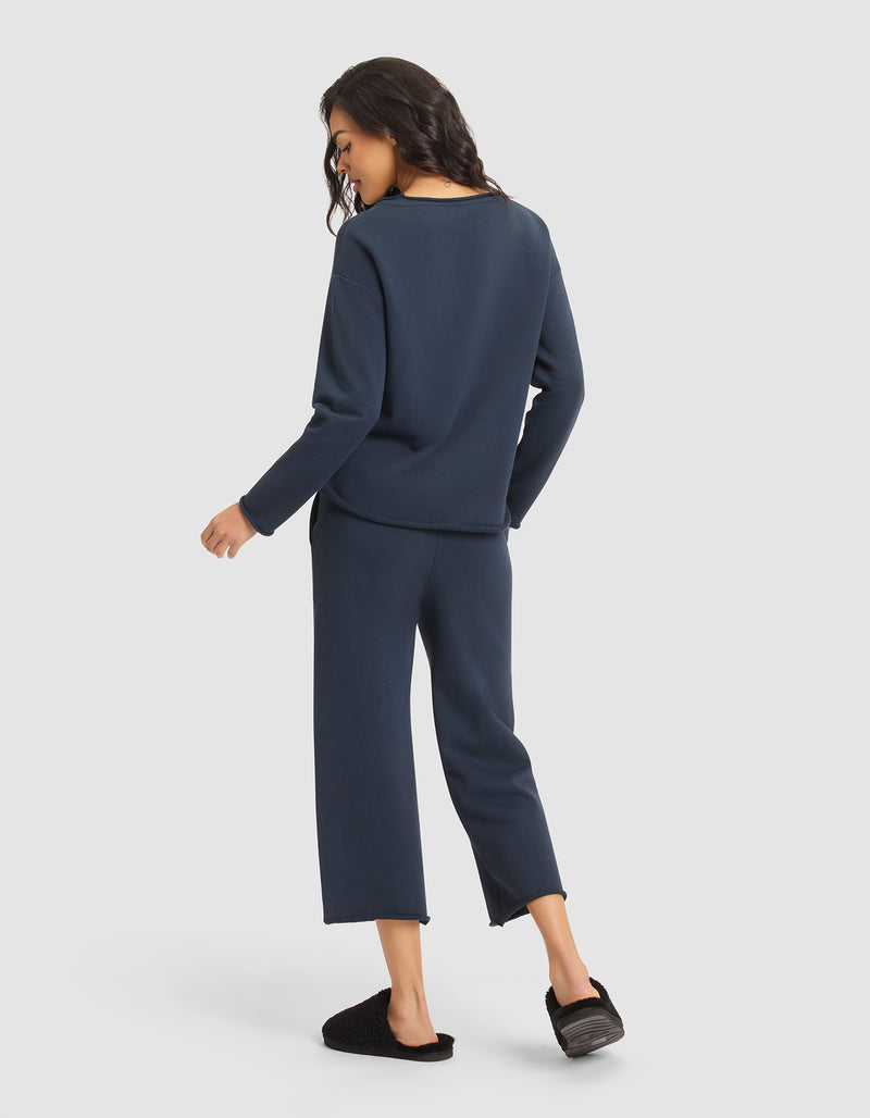 A woman models the Cozy Earth Women's Chelsea Set—dark blue long-sleeve top and matching wide-leg pants—with black slippers, standing against a light gray background.