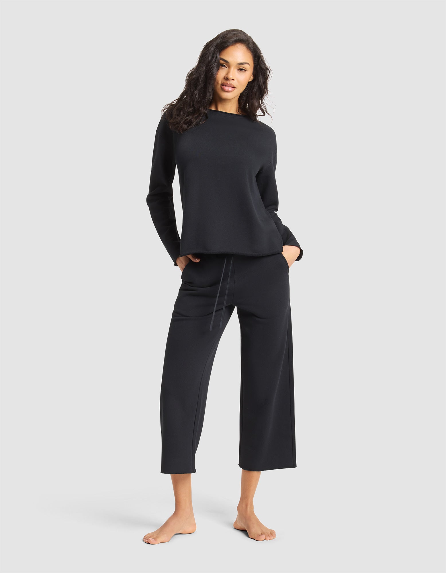 A woman with wavy dark hair models the Cozy Earth Women's Chelsea Set—matching black long-sleeve top and wide-leg cropped pants—standing barefoot with hands in her pockets against a plain light gray background. 