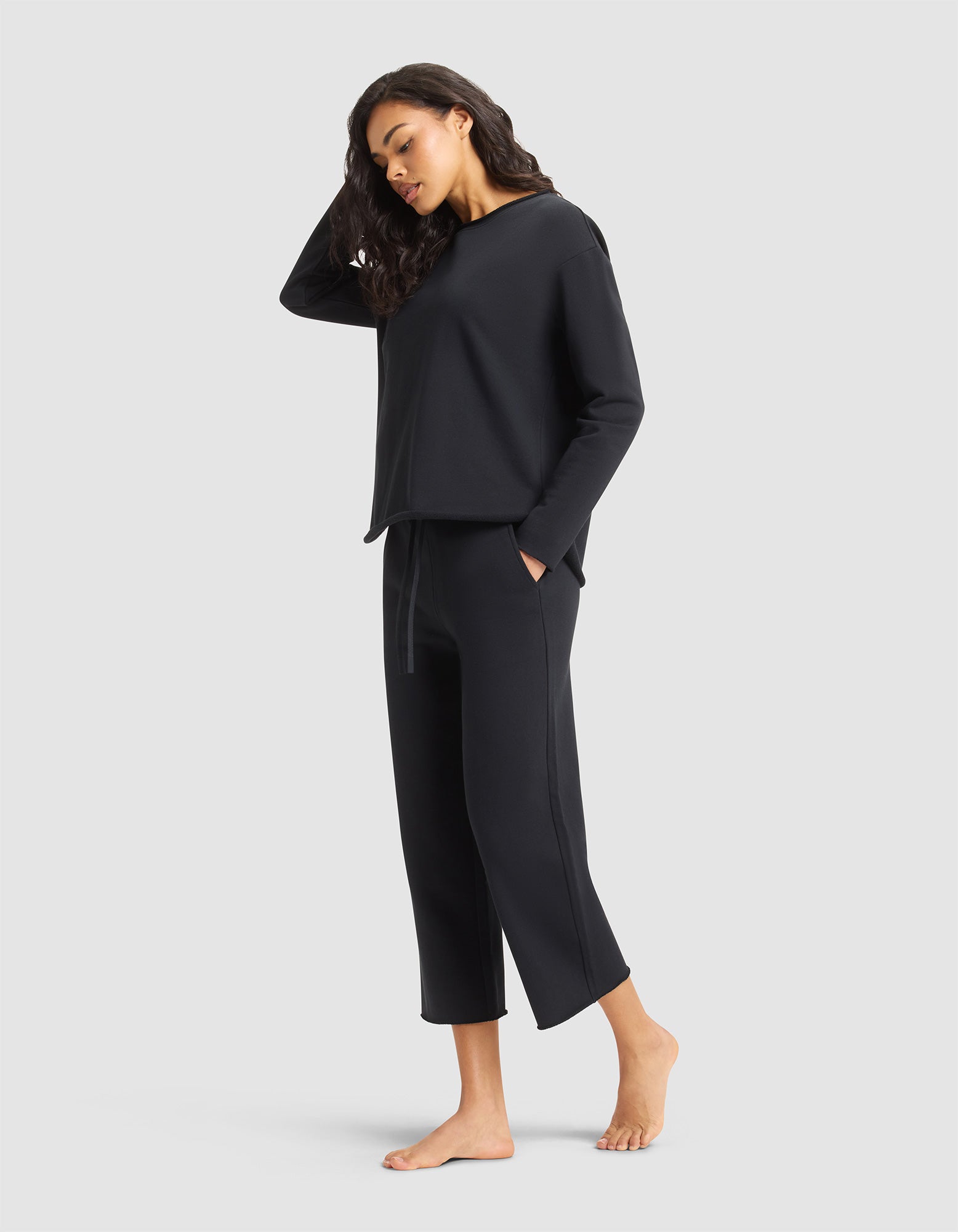 A woman with long dark hair stands barefoot against a light gray background, looking relaxed in the Cozy Earth Women's Chelsea Set—wearing a loose-fitting black top and matching cropped pants with her hands in her pockets. 