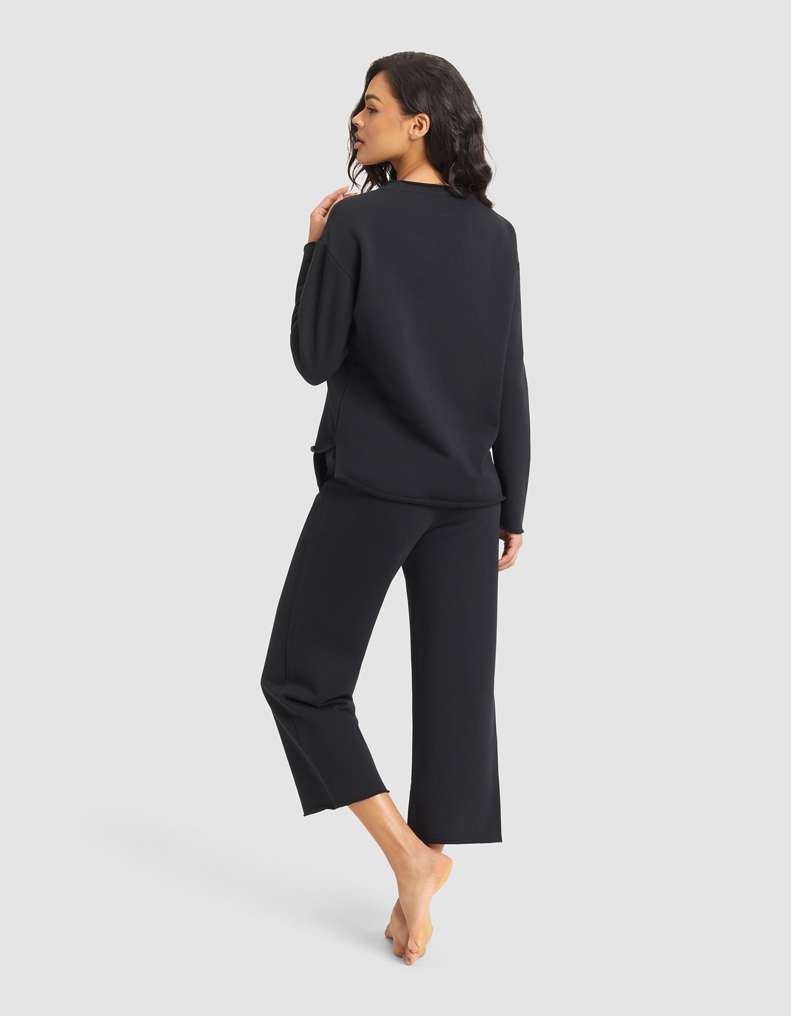 A woman stands barefoot, facing away, wearing the Cozy Earth Women's Chelsea Set—a loose-fitting black long-sleeve top and matching cropped pants—against a light background, her head turned to the side with her left hand on her shoulder. 