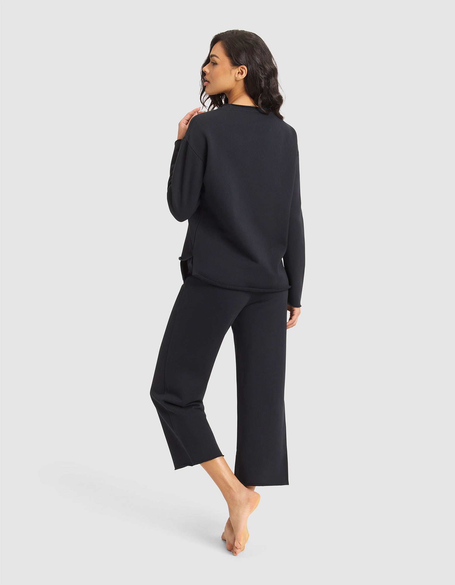 A woman stands barefoot, facing away, wearing the Cozy Earth Women's Chelsea Set—a loose-fitting black long-sleeve top and matching cropped pants—against a light background, her head turned to the side with her left hand on her shoulder. 