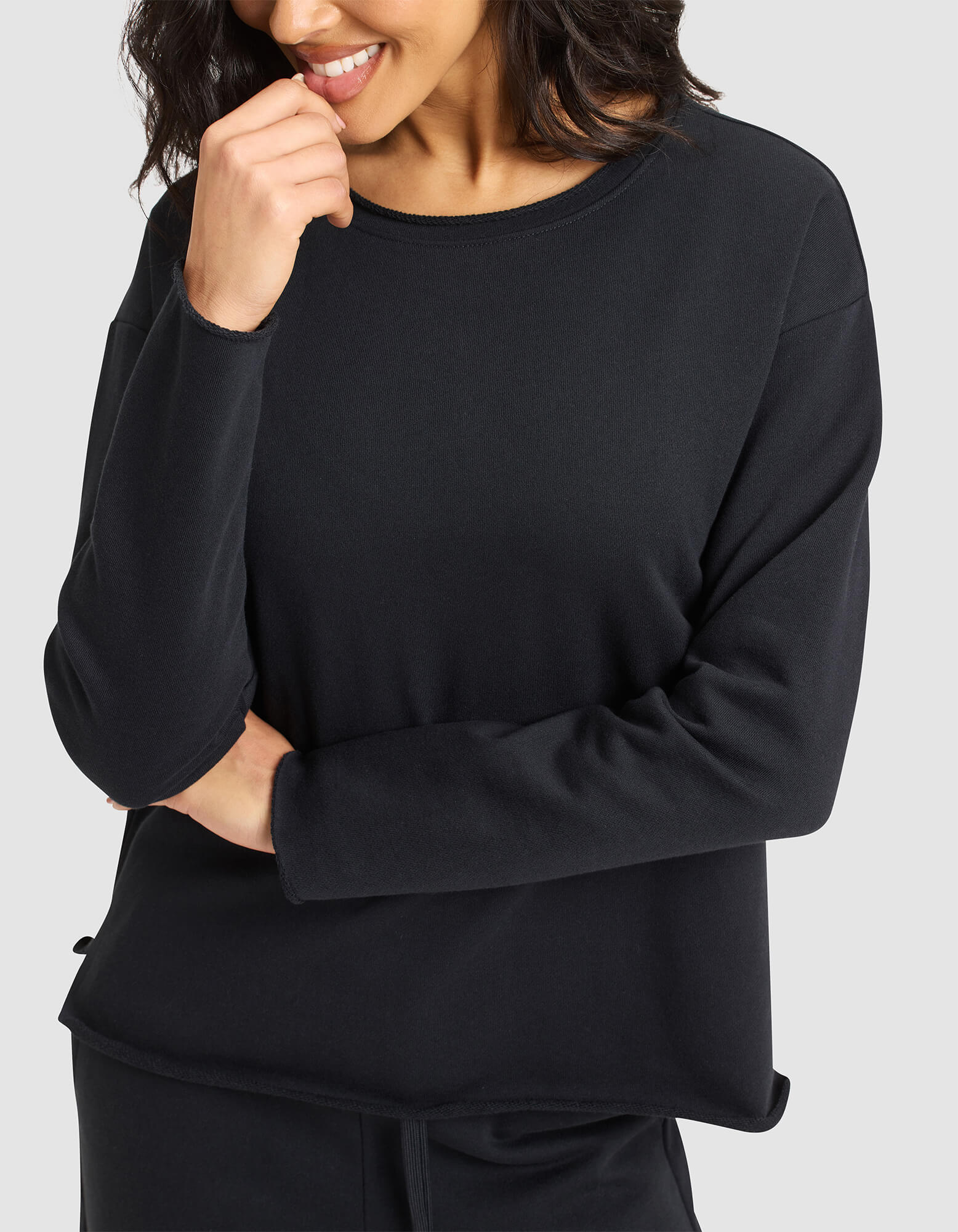 A woman with wavy dark hair smiles softly, holding her hand near her mouth while wearing Cozy Earth's loose-fitting Women's Chelsea Set in black. 