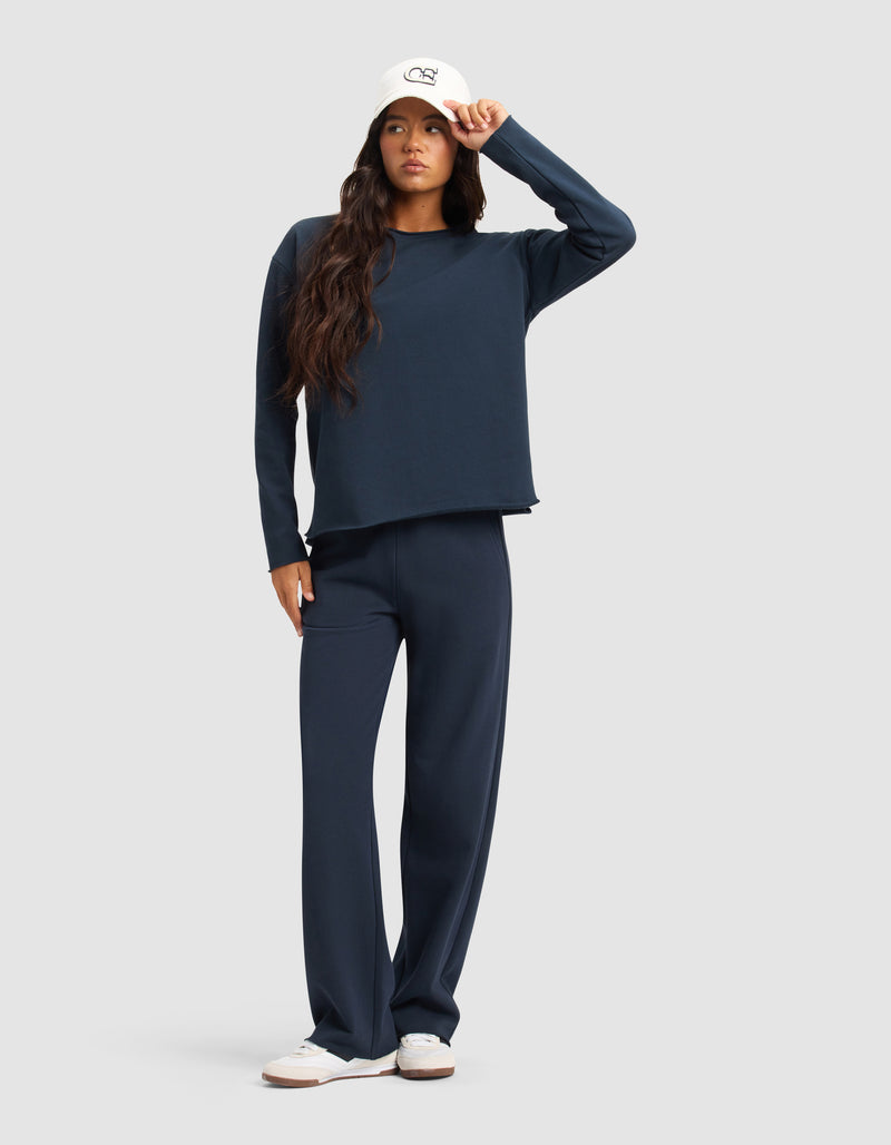 A woman models the Cozy Earth Women's Chelsea Set—navy long-sleeve top and wide-leg pants—paired with white sneakers and a baseball cap, standing against a plain light gray background and holding her cap’s brim.