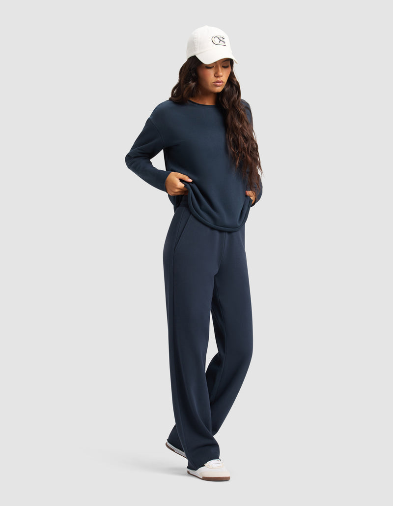 A woman wears Cozy Earth's Women's Chelsea Set in navy blue, featuring a long-sleeve top and wide-leg pants. She pairs it with a white baseball cap and sneakers, posing with hands in pockets against a light gray background.