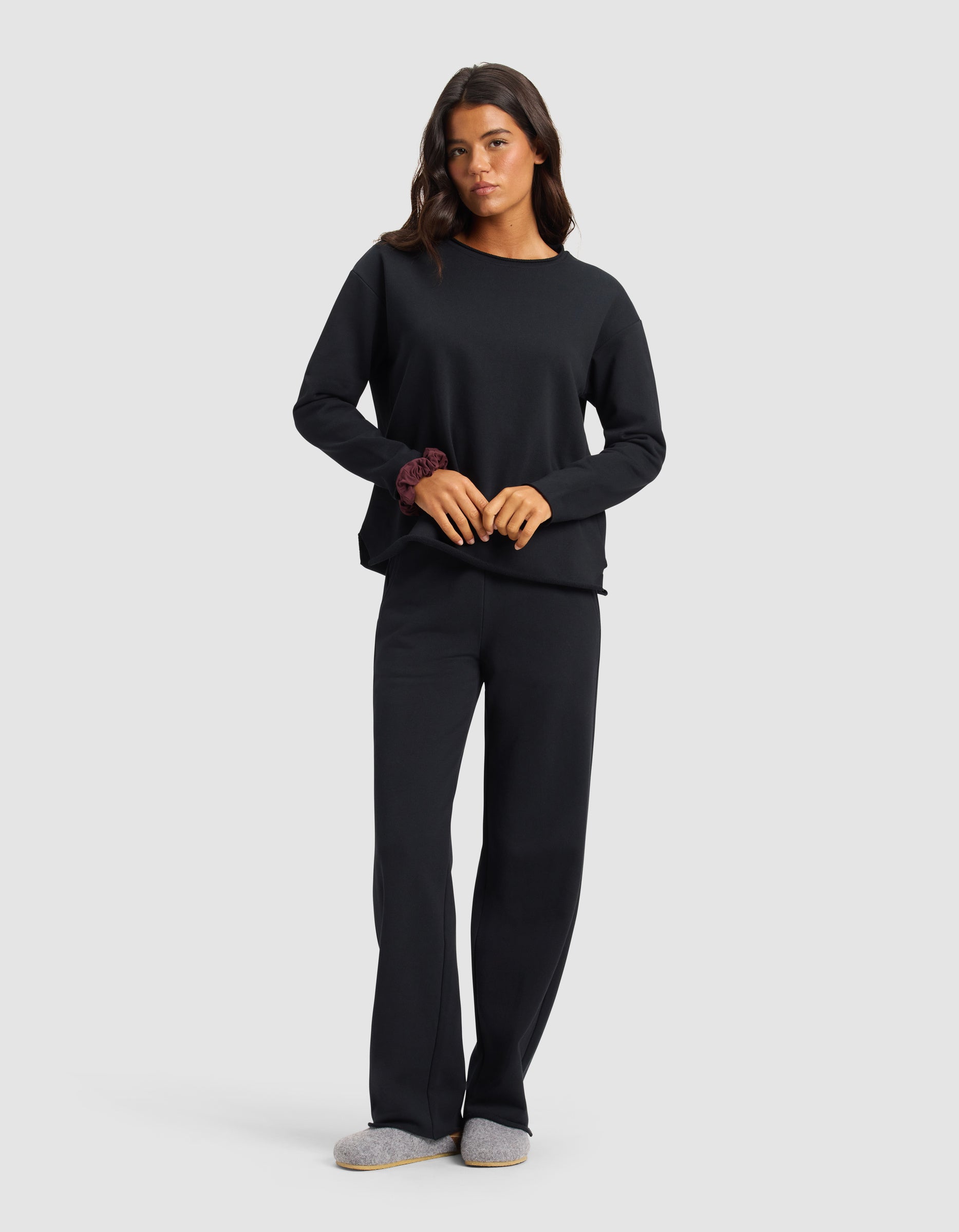 A woman wears the Women's Chelsea Set by Cozy Earth, featuring a loose-fitting black long-sleeve top and matching pants. She pairs the outfit with gray slippers, a scrunchie on her left wrist, and stands against a plain light background.