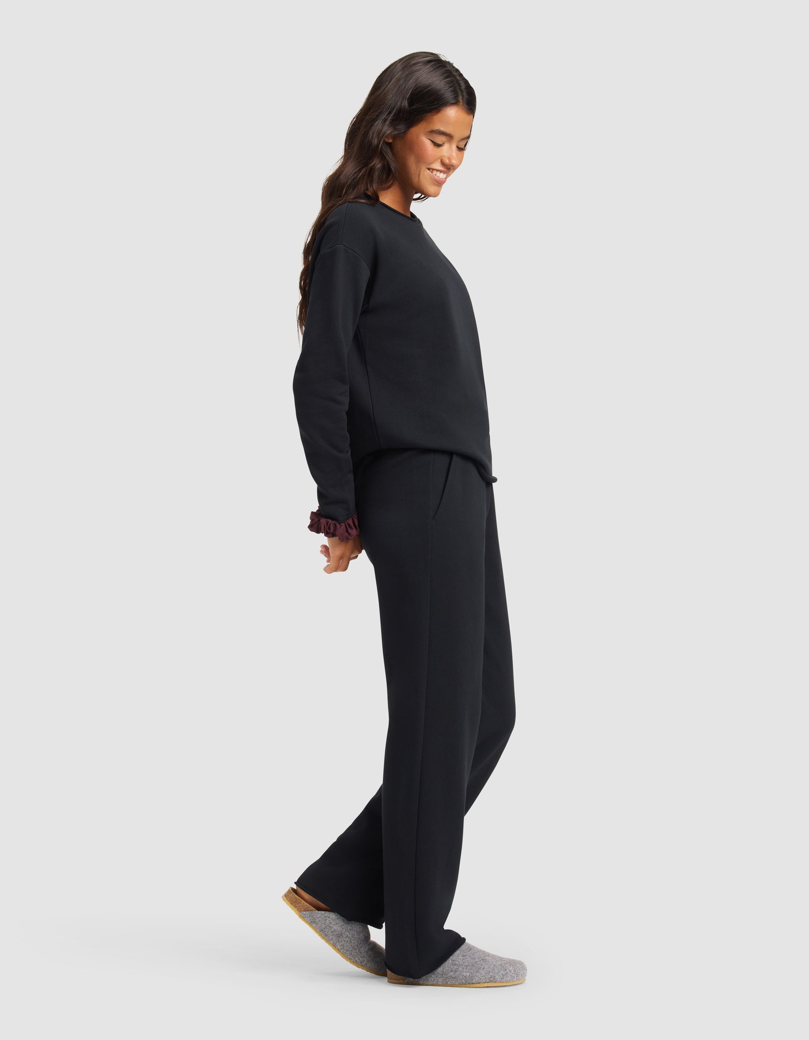 A woman with long dark hair smiles and looks down while wearing the Cozy Earth Women's Chelsea Set—loose black sweatshirt and matching wide-leg pants—standing against a plain light gray background.