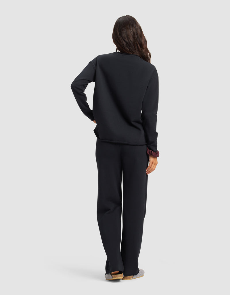 A person with long, wavy hair faces away, wearing the Cozy Earth Women's Chelsea Set—loose-fitting black sweatshirt and pants. One hand rests on their hip, slippers on their feet. The background is plain light gray.