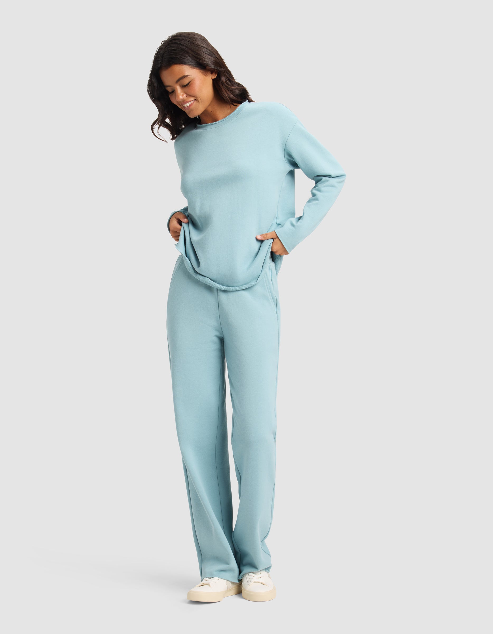 A woman smiles in the Cozy Earth Women's Chelsea Set—light blue long-sleeve top and matching loose pants with pockets—paired with white sneakers, set against a plain light background.|Color:River